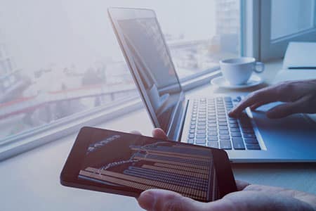 Laptop on a desk and a hand holding a phone displaying stock information.