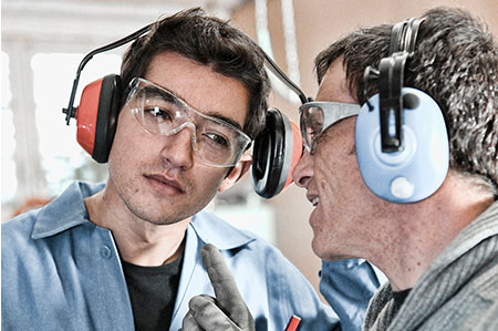 Zwei Arbeiter in der Fabrik tragen Gehörschutz versuchen im Maschinenlärm miteinander zu sprechen.