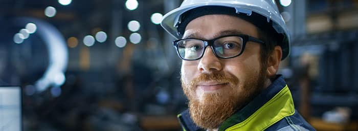 Uomo con indumenti ad alta visibilità e casco in una fabbrica che sorride alla telecamera.