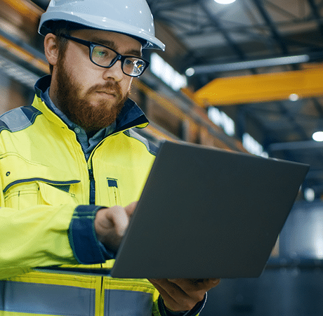 Mann in Warnkleidung und Helm in einer Fabrik bei der Arbeit am Computer.