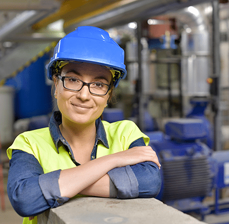 Frau in Warnkleidung und Helm in einer Fabrik, die in die Kamera lächelt.